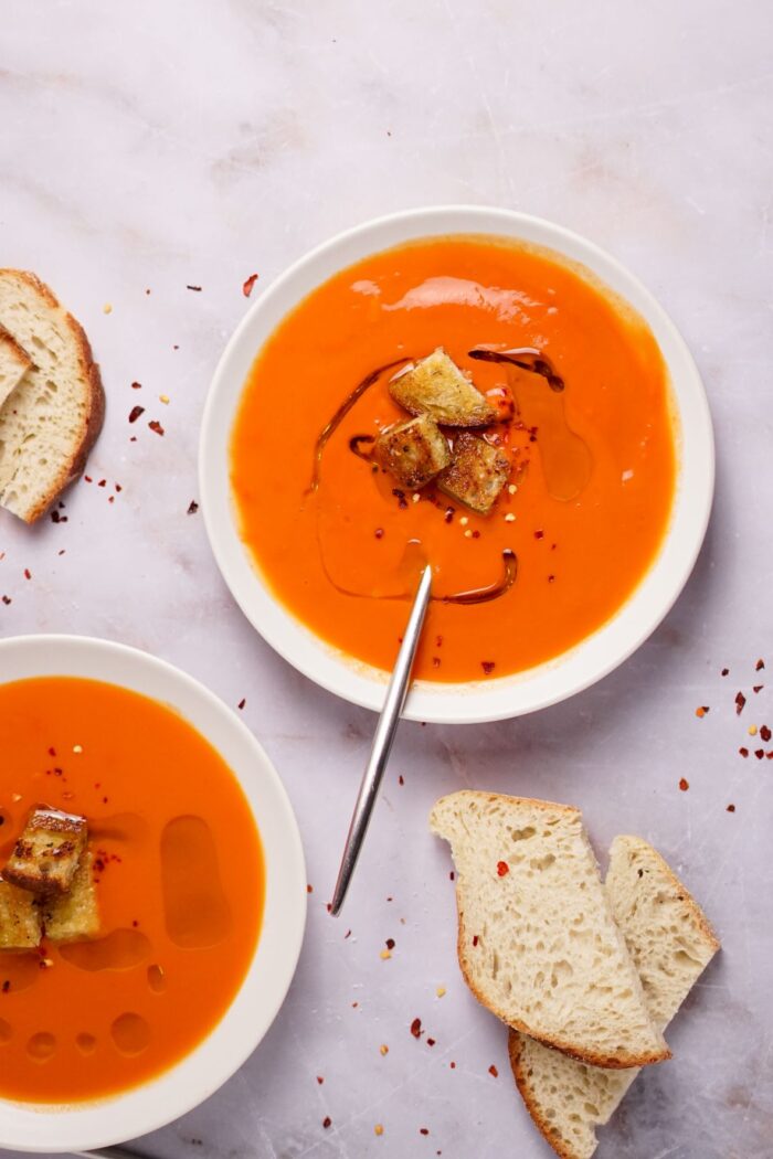 An overhead shot of two bowls of this healthy tomato soup recipe made croutons on top and sliced bread off to the side.